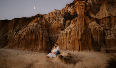 Pregnant couple sitting and snuggling in front of red sandstone cliffs with the moon rising behind them