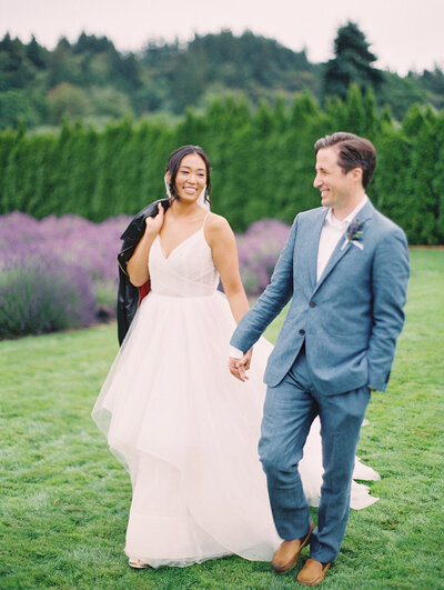 A smiling/laughing couple who are holding hands and walking on some grass in front of a blooming lavender field. The bride is holding a black leather jacket over her shoulder, and the groom is wearing a blue suit and leather moccasins and has his hand in his pocket.