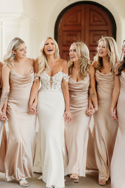 bride in a white wedding dress and bridesmaids in silk champagne dresses holding hands and laughing during relaxed bridal party portraits