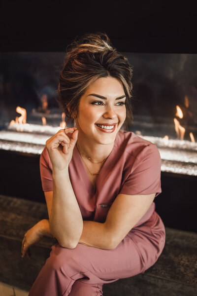 woman sitting in front of fireplace wearing pink scrubs while smiling with her hair in a bun