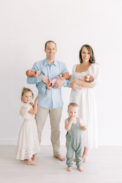 white studio with mom and dad holding triplet babies with two toddlers standing next to them.
