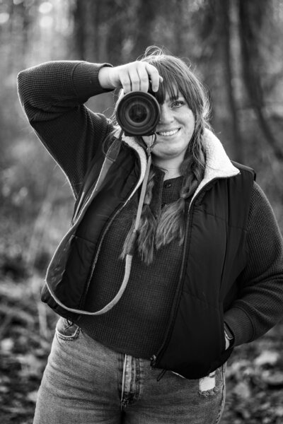 Black and white shot of Savannah holding her camera, smiling playfully, as warm sunlight creates a soft, radiant glow behind her.