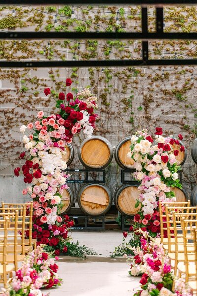 A split floral arch filled with red, white, and pink roses sits in front of a row of wine barrels on an ivy-covered patio. Gold chairs are placed in rows with a center aisle for the wedding processional.