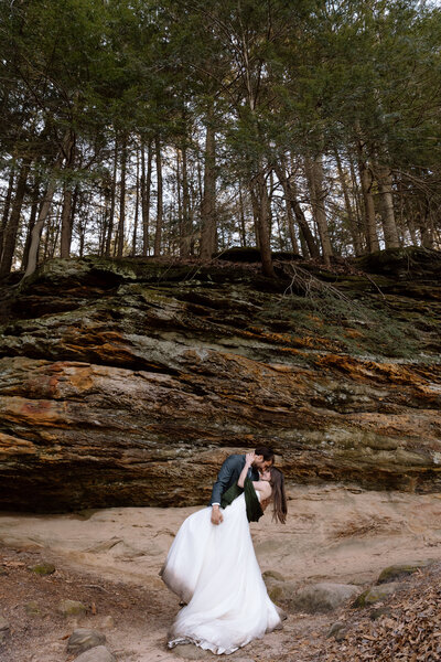 Groom dipping bride, a cliff and trees are behind them
