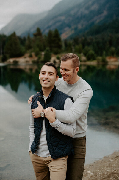 A couple shares a tender moment by a mountain lake, surrounded by lush greenery.