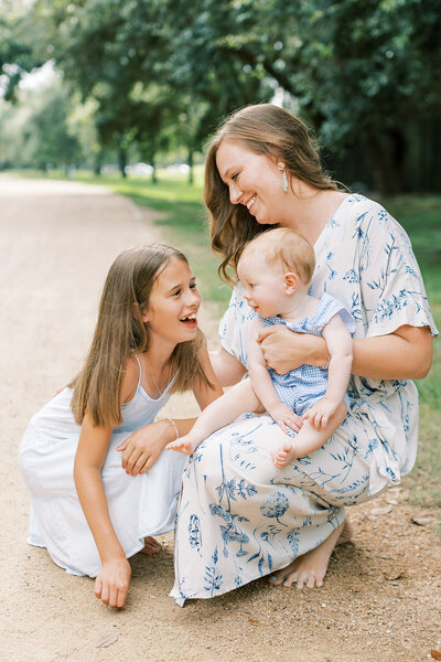 Mom in blue floral dress kneeling on the ground with her daughter and holding a baby on her leg