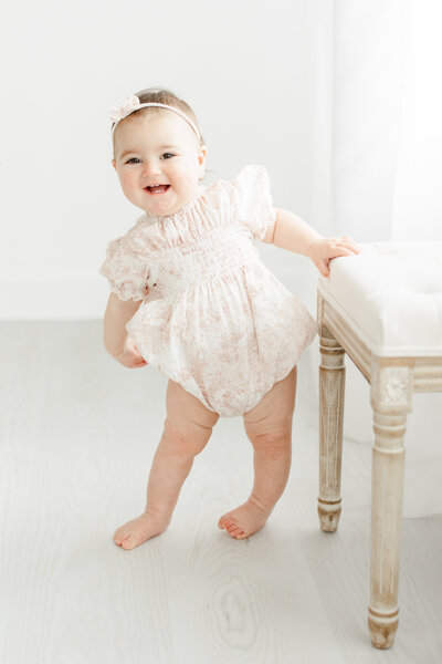 One year old baby girl stands off kilter next to chair and smiles during first birthday portrait session