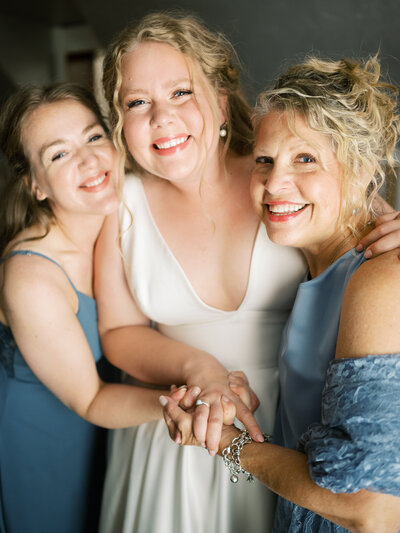 A bride smiles as her mom and sister hug her while she's getting ready for her wedding.