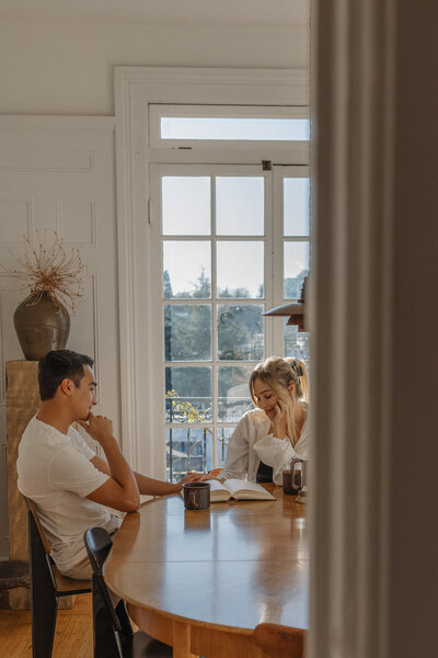 A man and woman sit at a wooden dining table with coffee mugs and an open book between them, sharing a quiet moment – Family Autism Support Coach at Chantal Hewitt Coaching