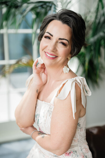 Kari Dawson, lead wedding photographer of Kari Dawson + Co, seated in soft natural light wearing a floral dress, smiling gently with hand resting near her cheek, exuding warmth and calm professionalism