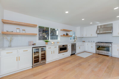 Modern white kitchen remodel by AW Development featuring shaker cabinets, open wood shelving, gold hardware, stainless steel appliances, and wide plank flooring.