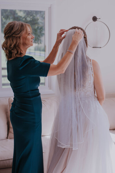 A woman in a dark green dress adjusts the veil of a bride in a white gown at Halcyon Watson. They are in a softly lit room with a window, revealing greenery outside. A cream-colored couch and an elegant wall-mounted light fixture add charm to the setting.
