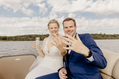 A couple in wedding attire sits on a boat, smiling as they display their rings. The bride holds a champagne flute, and the serene lake backdrop glimmers under a partly cloudy sky—captured beautifully by Brainerd Lakes Area Photographer.