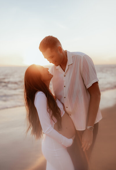 Expecting couple standing close to each other on the beach at golden hour. 