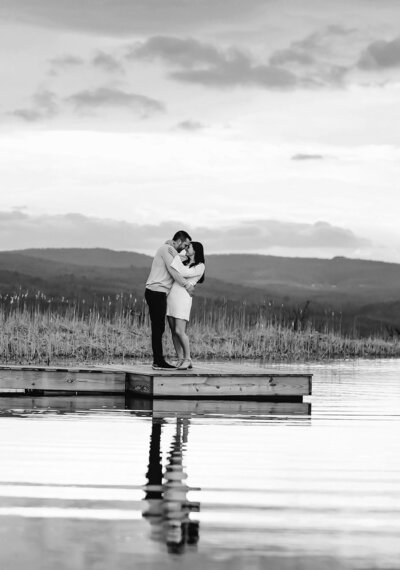 Romantic black and white engagement photo of a couple embracing on a dock, surrounded by water and distant mountain views.