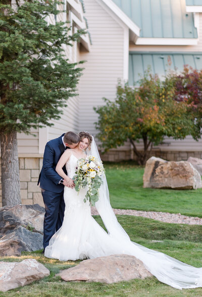 denver wedding photographer captures bride and groom posing outside of their Denver wedding venue with the groom leaning in and kissing his brides neck as the bride holds a large wedding bouquet photographed by Denver-wedding-photographer-micro-wedding-photographer