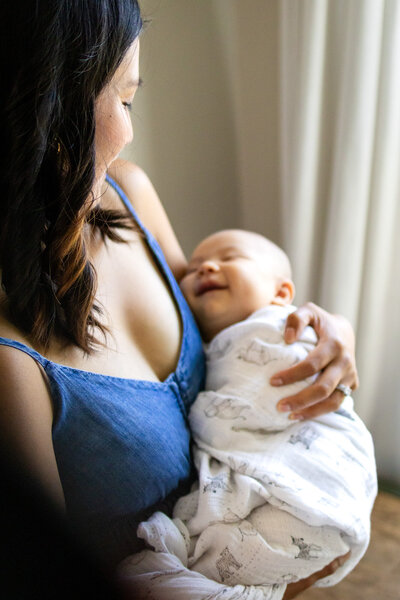 A mama holding her swaddled baby as he smiles up at her, captured in soft indoor light – ellobelle photography.