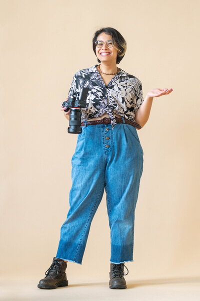 Erin Deleon, slightly androgynous fem Filipino photographer holding her camera and posing in front of a beige backdrop