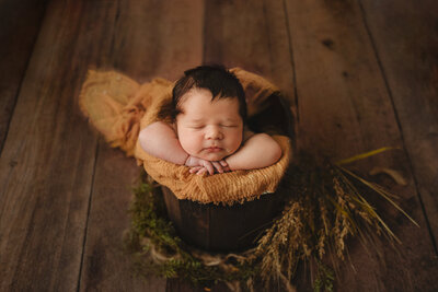 baby boy in a wooden bucket with his head on his hands and a faded mustard wrap under his hands