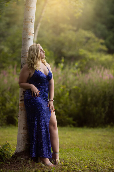 High school female leanign on a birch tree in Leominster wearing her blue sparkling prom dress with a purple flowers and greenery in background