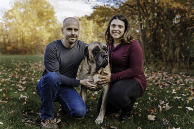 Verona B Photography captures a family and their dog at Silver Creek Metro Park in Norton, Ohio. The couple is kneeling down and have their hands on the dog while smiling at the camera with beautiful fall foliage in the background. 