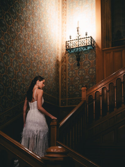 A woman in a white fringe dress ascends an ornate wooden staircase with vintage wallpaper in warm lighting.