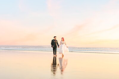 bride and groom walking along the beach with their reflections showing on the wet sand