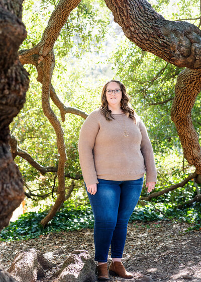 Woman in a tan sweater and jeans standing outdoors surrounded by large twisted tree branches and sunlight filtering through the leaves.