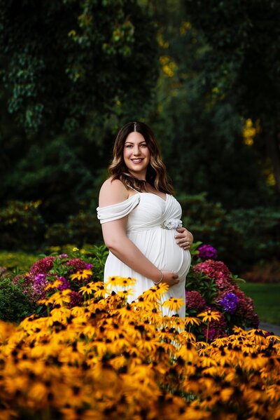 Pregnant woman in a white dress surrounded by yellow flowers at Deer Lake Park in Burnaby during an outdoor maternity photoshoot.
