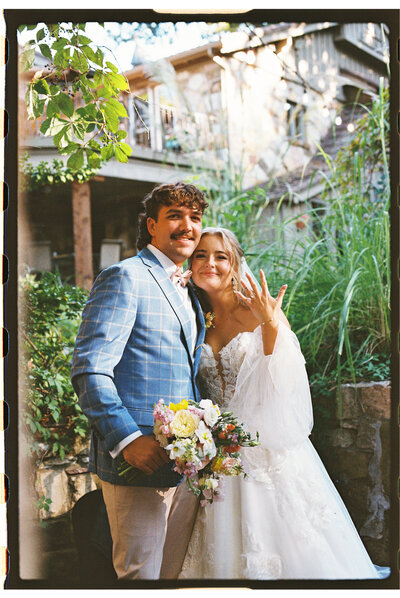 A bride in a white gown and a groom in a blue checkered jacket hold hands around a stone corner of a rustic building. Shes holding a bouquet, both surrounded by greenery, with sunlight filtering through leaves.