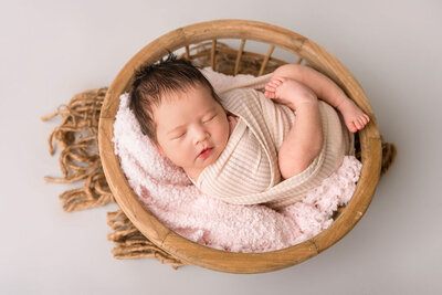 Professional Connecticut newborn photographer specializing in posed newborn portraits featuring baby wrapped in beige in wooden bowl with pink textured layers