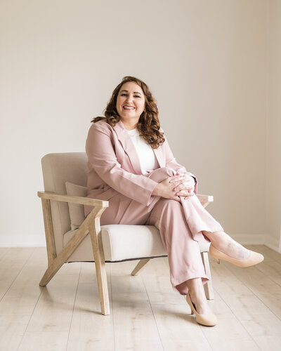 Dark brunette woman sitting in a chair with legs crossed, wearing light pink pants and matching dress jacket with a white shirt under the jacket, smiling at the camera