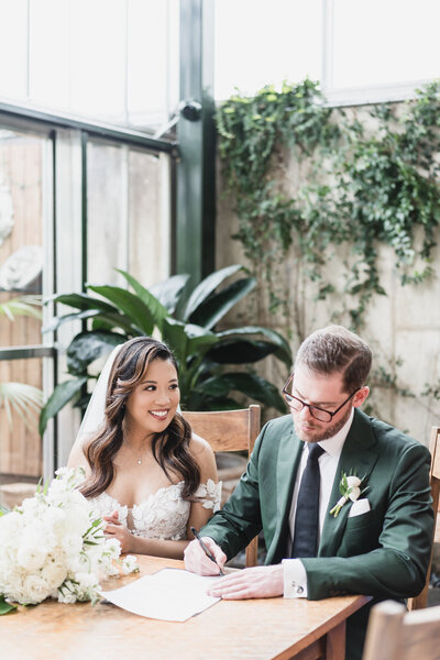 Bride and groom smile while signing marriage license in a lush greenhouse venue at Planterra Conservatory, West Bloomfield Michigan