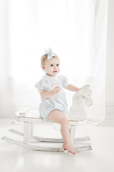Little girl in blue romper sits on a white rocking horse