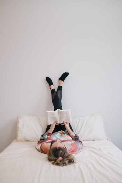 Sammy laying on a bed reading a book with her legs on the wall