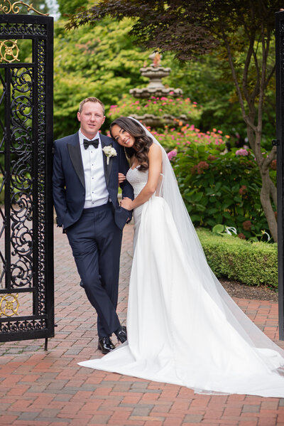 A bride wearing a long white gown holding onto her groom who is wearing a black and navy tuxedo outside their wedding at the Ashford Estate in Allentown NJ.  He is leaning on a black wrought iron decorative gate and you can see a flower filled fountain in the garden behind them.