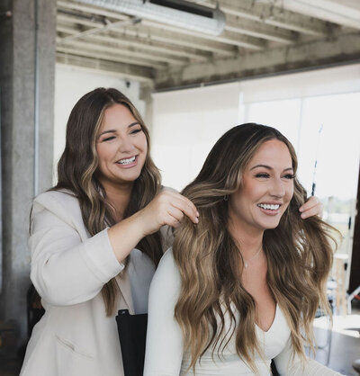 A hairstylist is smiling and facing a mirror with her cient, showcasing the end result of her color and hair extensions transformation.