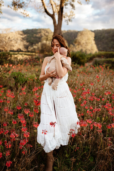 A woman wearing a white dress stands in a field of red flowers, holding a baby in her arms. Trees and hills are visible in the background.