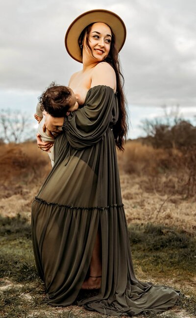 Woman in a flowing green dress and hat breastfeeds a child while standing in an open field.