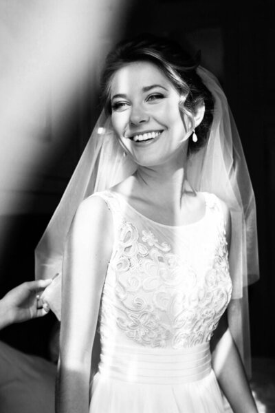 A bride smiles in the light as a brides maid helpers her get ready.