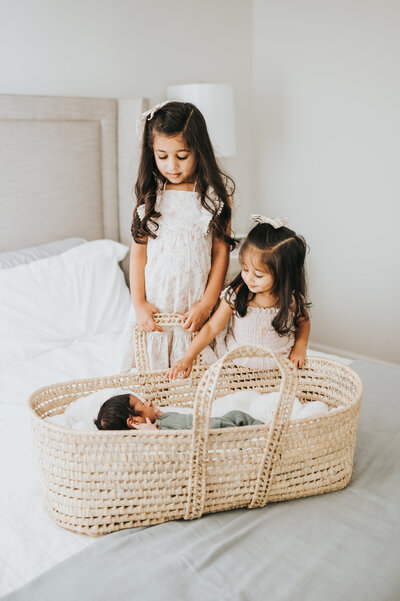 Two big sisters look down at newborn baby boy in moses basket during in-home photography newborn session