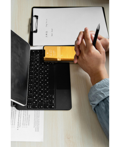 A person hold their own hands in front of a iPad, phone and clip board