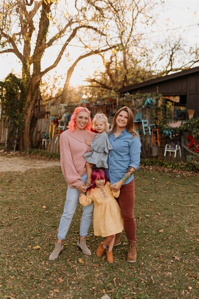 Lesbian couple hold each other and their two daughters in front of tree