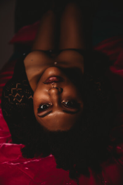 Beautiful Black Woman Wearing Black Body Suit and Laying on Hot Pink Bedding, Making Fierce Eye Contact with Camera