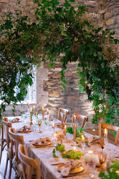 Long reception table styled with floral centerpieces, taper candles, and custom menus in a warm-toned, romantic setting.