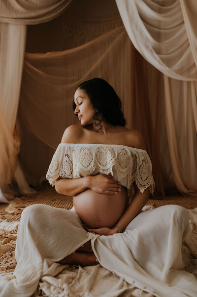 A bare belly pregnant woman sitting on the floor of a fort worth texas photography studio holding her belly with both arms