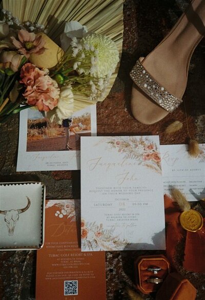 A flat lay of wedding details from a Tubac Golf Resort wedding in Tucson, Arizona, including a floral bouquet, wedding invitations, and a pair of embellished sandals.