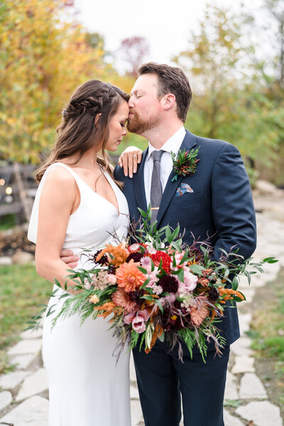 A bride rests her hand on her groom's shoulder while he kisses her forehead, surrounded by fall colors