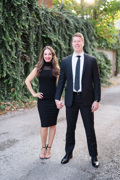 A young couple in black formal attire hold hands and smile during their German Village engagement