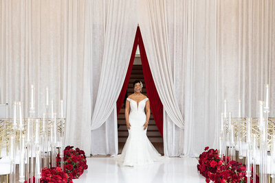 Bride in an off-the-shoulder lace gown walking down a white aisle lined with red roses and tall glass candle holders, framed by dramatic white draping and bold red curtains, designed by Wells Design Co.